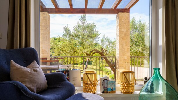 A room with a blue armchair, a small table, and a view of a balcony with a railing and greenery.