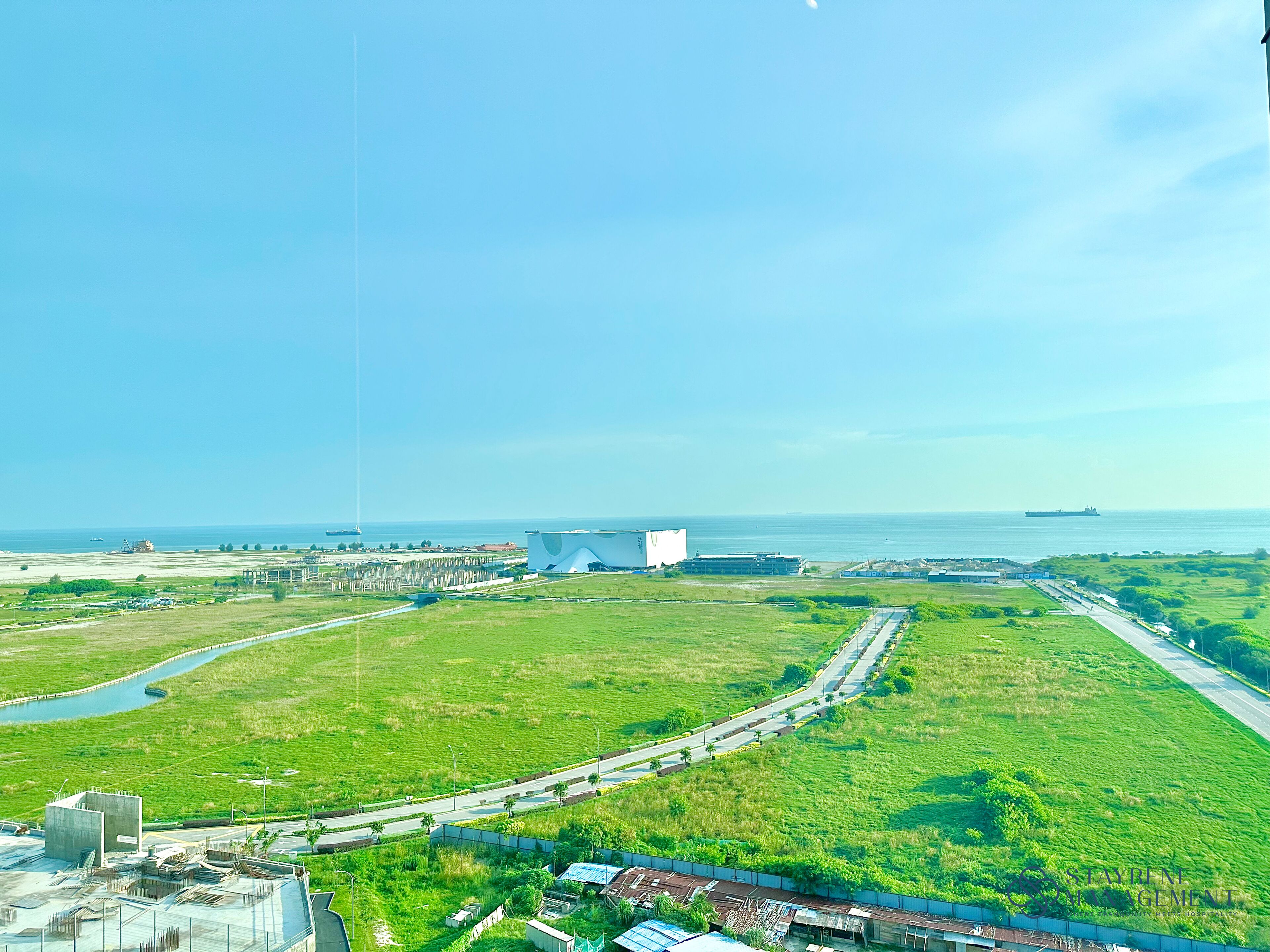 An aerial view of a coastal area with a large white building, green fields, and a road leading to the sea.