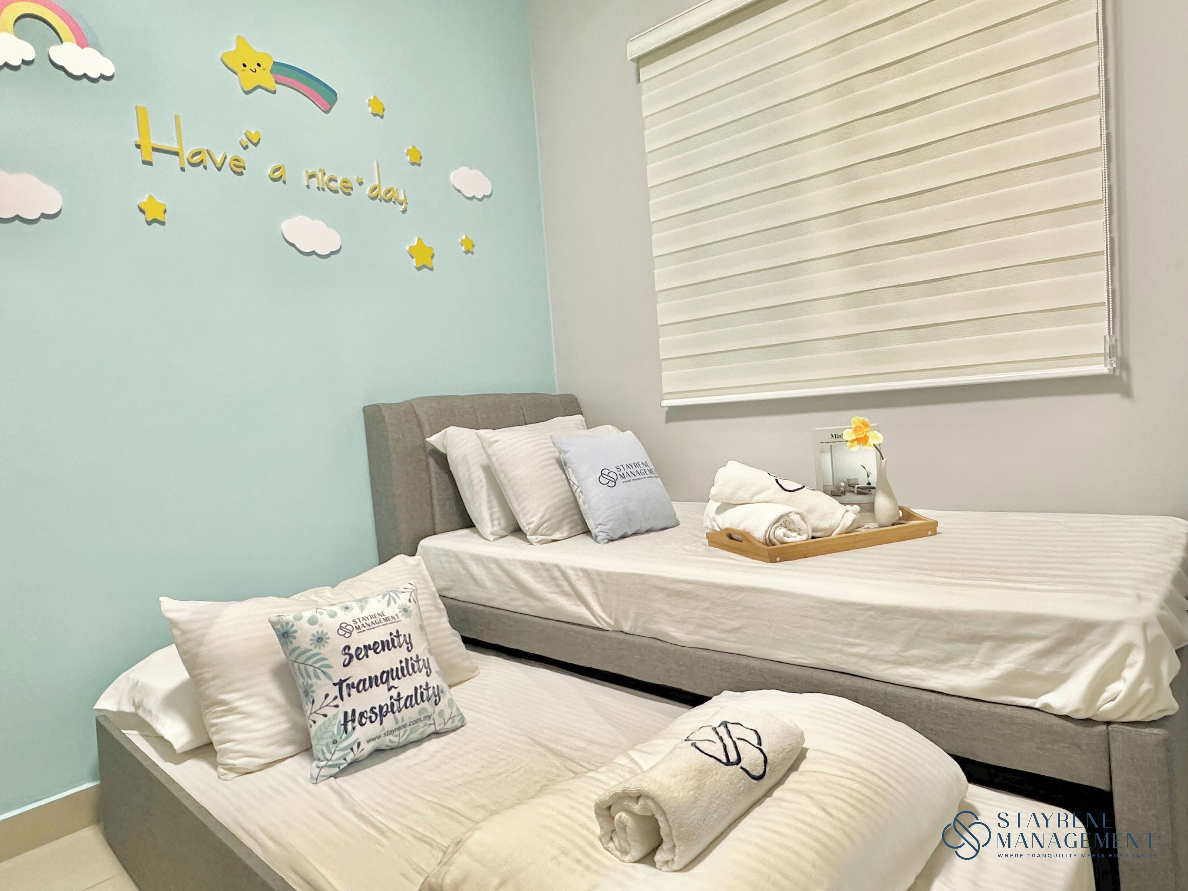 A hotel room with two beds, a window with blinds, a wall with a 'Have a nice day' message, and a tray with towels and a vase with a flower.