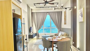 A modern living room with a dining area, a ceiling fan, and a large window with a city view. - Amber Cove Melaka Sea View by Stayrene (Malacca City)