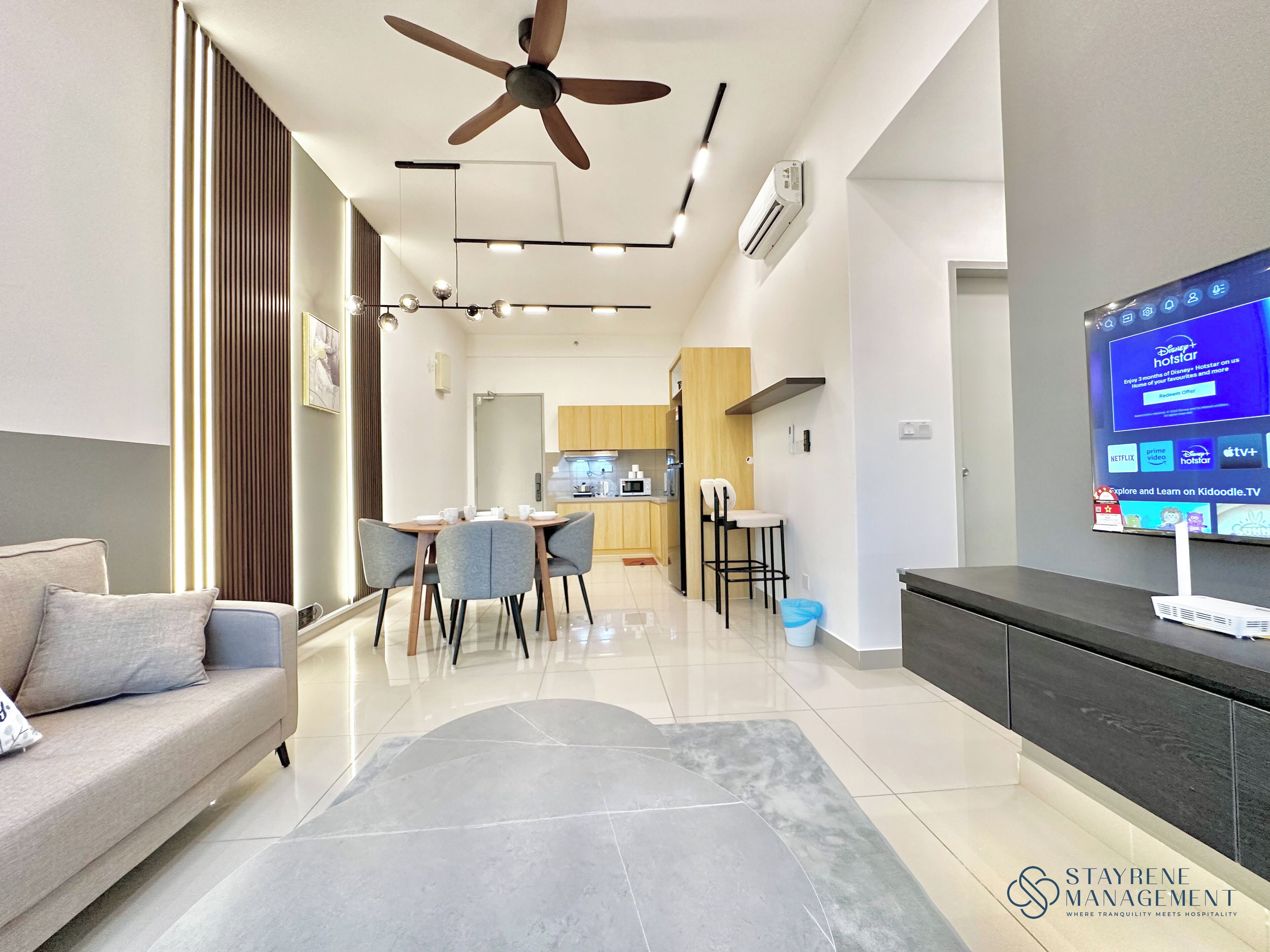 Una sala moderna con un sofá, un comedor con sillas, una cocina al fondo y un televisor que muestra Disney+ Hotstar.