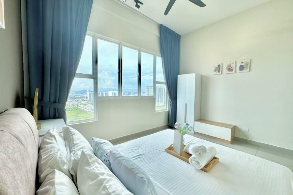 Una habitación de hotel moderna con una cama grande, una mesita de noche con un florero y vistas a la ciudad a través de amplios ventanales.