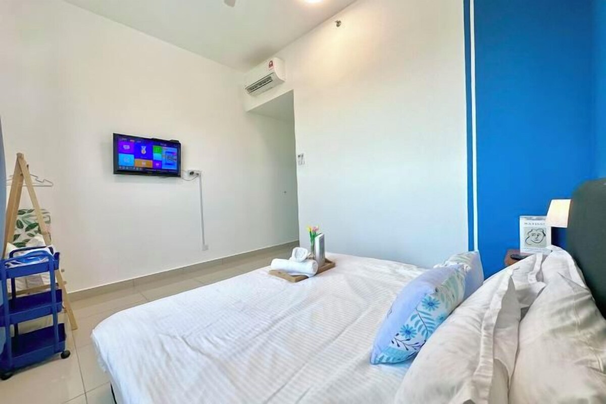 Habitación de hotel con cama, televisor en la pared, una mesita con un florero y un libro, y un carrito azul.