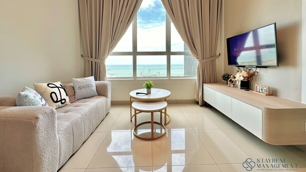 A modern living room with a sofa, a small round table, a TV mounted on the wall, and a view of the sea through large windows. - Amber Cove Melaka Sea View by Stayrene (Malacca City)
