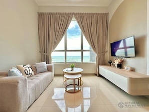A modern living room with a sofa, a small round table, a TV mounted on the wall, and a view of the sea through large windows. - Amber Cove Melaka Sea View by Stayrene (Malacca City)