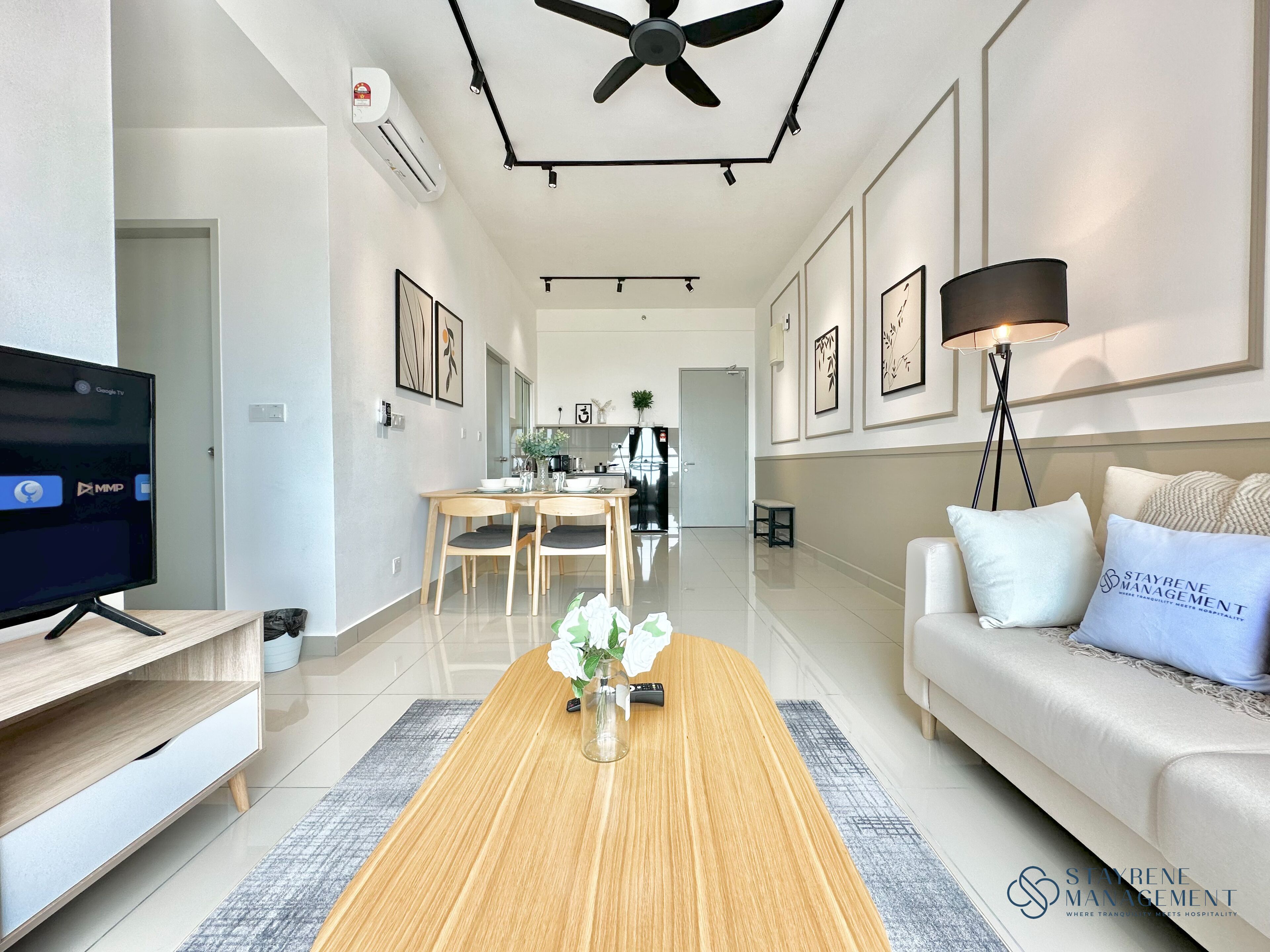 A modern living room with a flat-screen TV, a wooden coffee table with a vase of flowers, a sofa with branded pillows, and a dining area with a table and chairs.