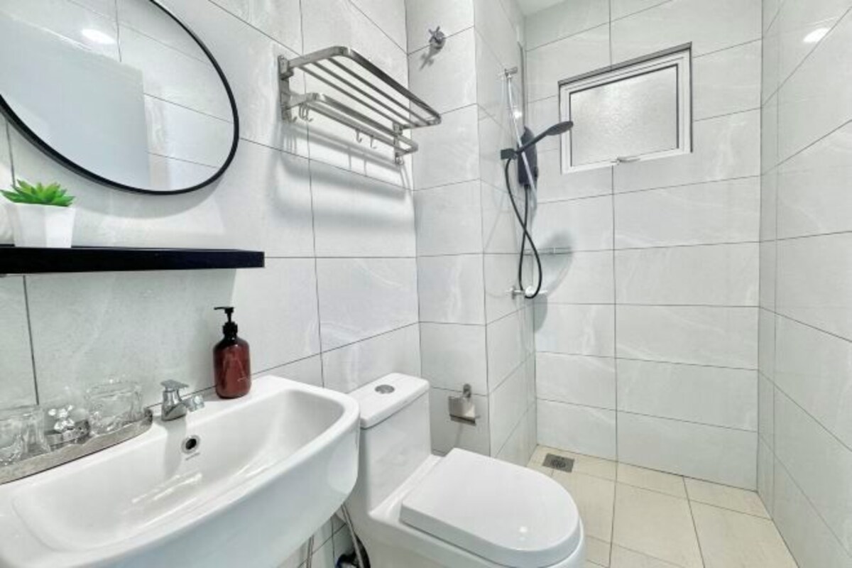 Un baño moderno con lavabo, inodoro y ducha blancos. Hay un espejo sobre el lavabo y un perchero en la pared.