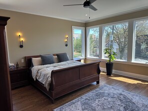 A bedroom with a large bed, two bedside tables, a ceiling fan, and a view of the outside through large windows.