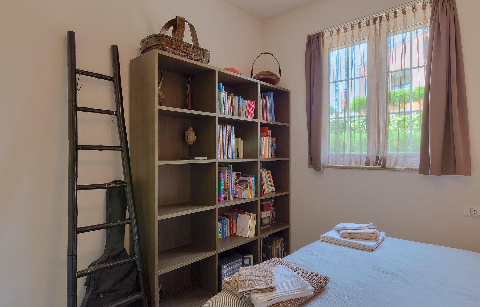 A bedroom with a bed, a bookshelf, a ladder, and a window with curtains.