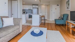A modern living room with a sofa, a coffee table, and a TV. There is a kitchen area in the background with cabinets and a refrigerator.