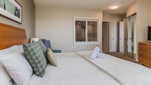 A hotel room with a bed, a wooden headboard, a bedside table, a dresser, a mirror, and a window with blinds.
