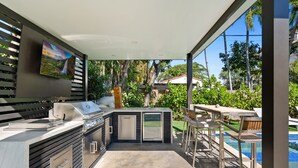Een buitenkeuken met een barbecue, een televisie erboven en uitzicht op een zwembad en groen.