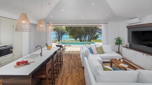 Et moderne køkken med en central ø, et spiseområde med udsigt til poolen, og en opholdsstue med sofa og TV.