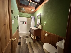 A bathroom with a wooden floor, green walls, a white bathtub, a wooden vanity with a mirror, and a toilet. - Cozy 2br Bear Den Cabin in Campobello SC, on the upcoming Saluda Grade Trail. (Campobello)
