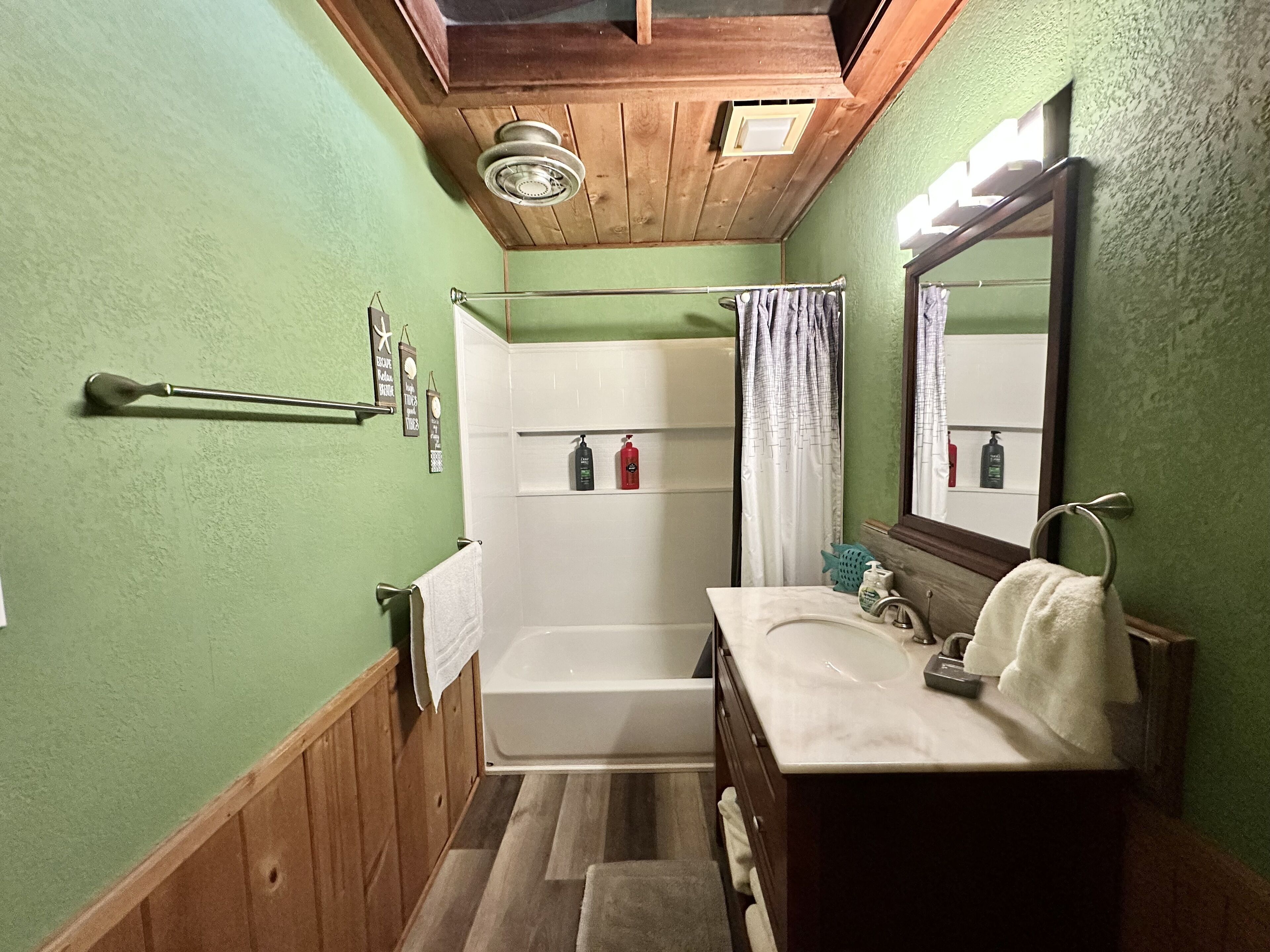 Une salle de bain avec un plafond en bois, des murs verts, une baignoire blanche, un lavabo avec un miroir et un rideau de douche.