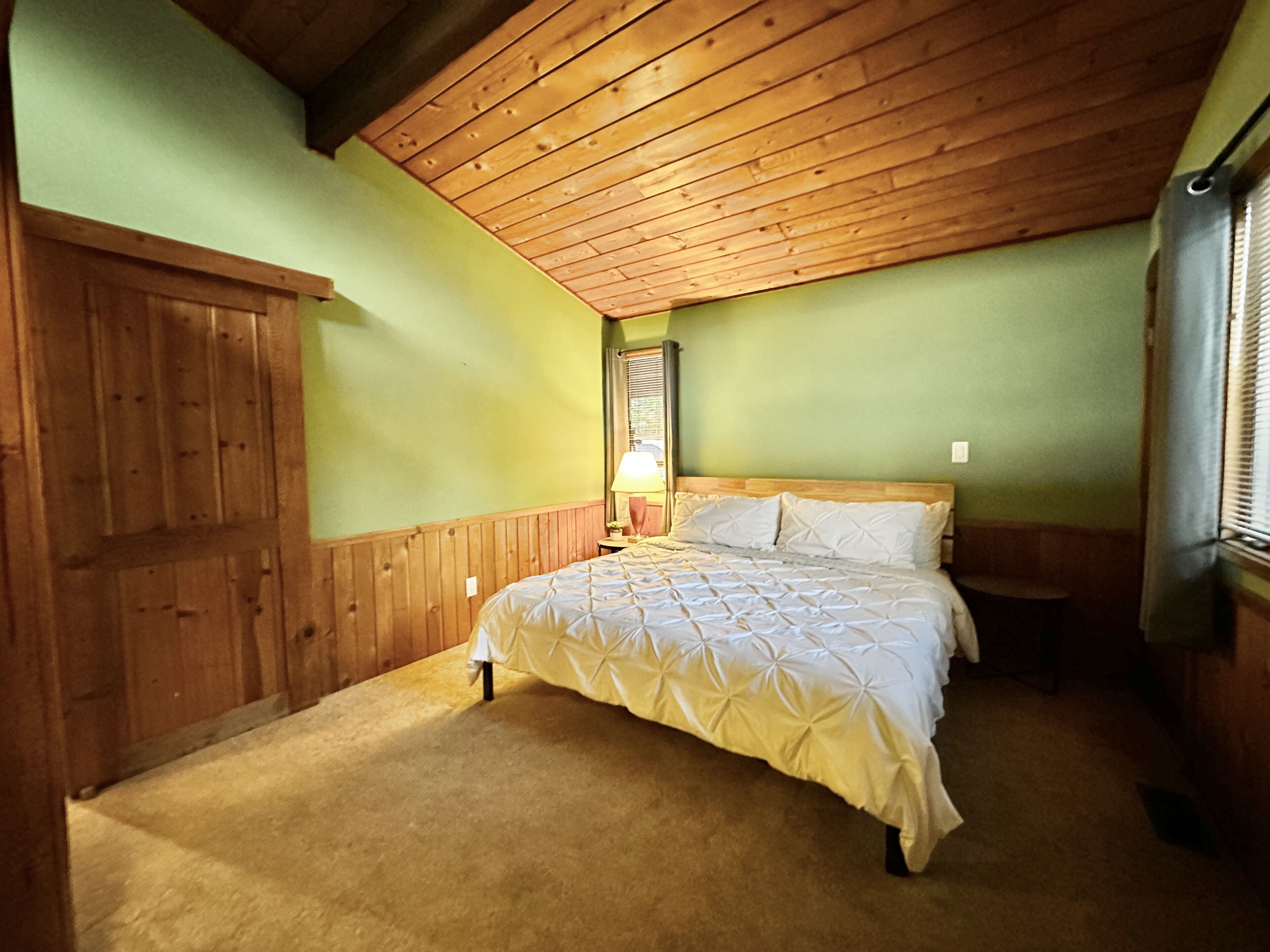 A bedroom with a wooden ceiling, a bed with a quilted cover, a nightstand, and a window with blinds.