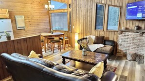 A cozy room with a wooden interior, a leather sofa, a dining table with chairs, a TV mounted on the wall, and a fireplace. - Cozy 2br Bear Den Cabin in Campobello SC, on the upcoming Saluda Grade Trail. (Campobello)