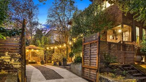 A well-lit garden area with a pathway leading to a wooden gate, surrounded by various plants and trees. - Artistic Victorian Gem in a Trendy neighborhood (Portland)