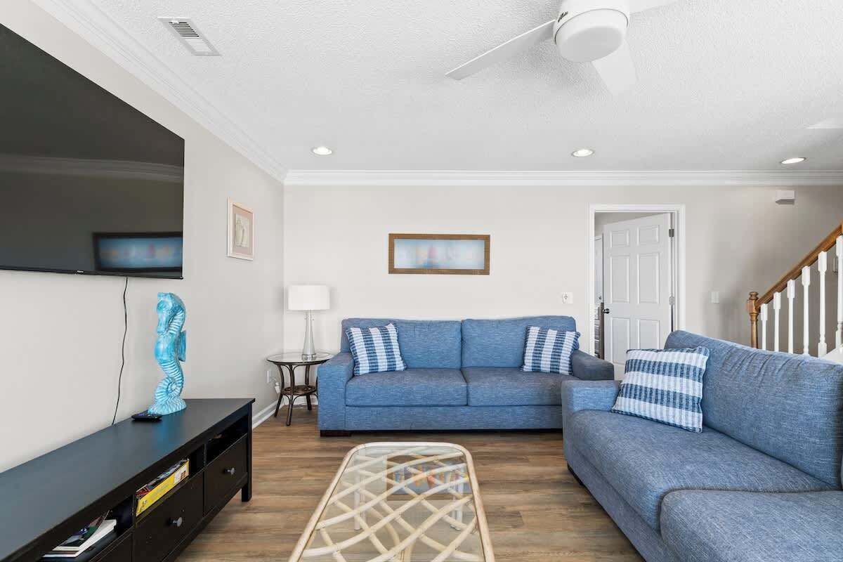 A living room with a blue sofa, a flat-screen TV, a ceiling fan, and a staircase.