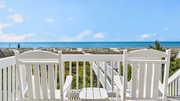 A wooden deck with white chairs and a table, overlooking a beach with clear blue water and a distant shoreline. - Zion of Holden by Brunswick Vacation Rentals: 5BR Oceanfront w/ Pool & Views (Holden Beach)