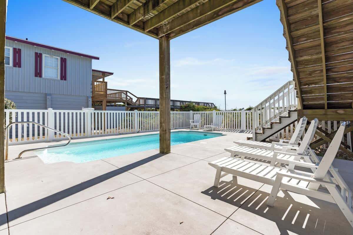A pool area with a wooden deck, white lounge chairs, and a white fence.
