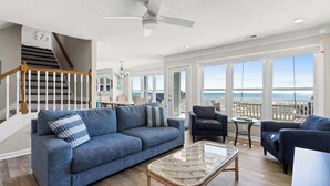 A spacious living room with a blue sofa, a glass coffee table, and a view of the ocean through large windows. - Zion of Holden by Brunswick Vacation Rentals: 5BR Oceanfront w/ Pool & Views (Holden Beach)