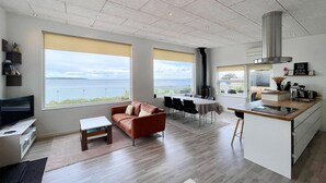 Una sala de estar moderna con un sofá, una mesa de centro y un televisor. Hay un comedor con una mesa y sillas, y una cocina con estufa y cafetera. Amplias ventanas ofrecen vistas al mar.