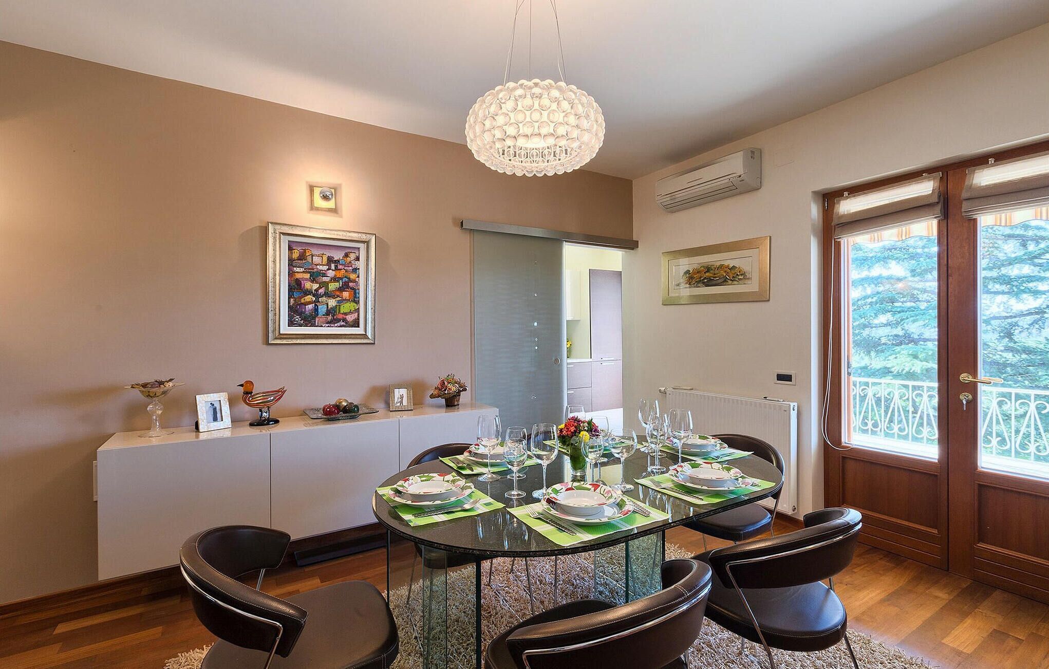 A dining area with a round glass table set for six, surrounded by black chairs. There is a chandelier above, a framed picture on the wall, and a window with a view of trees.