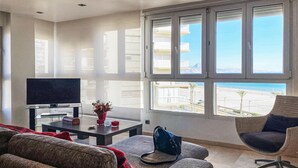 Ein Wohnzimmer mit Fernseher, Couchtisch mit Dekoration, Sofa, Sessel und großen Fenstern mit Blick auf den Strand.