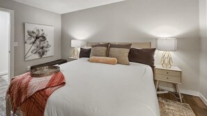A bedroom with a large bed, two bedside tables with lamps, a wall-mounted artwork, and a red throw blanket.