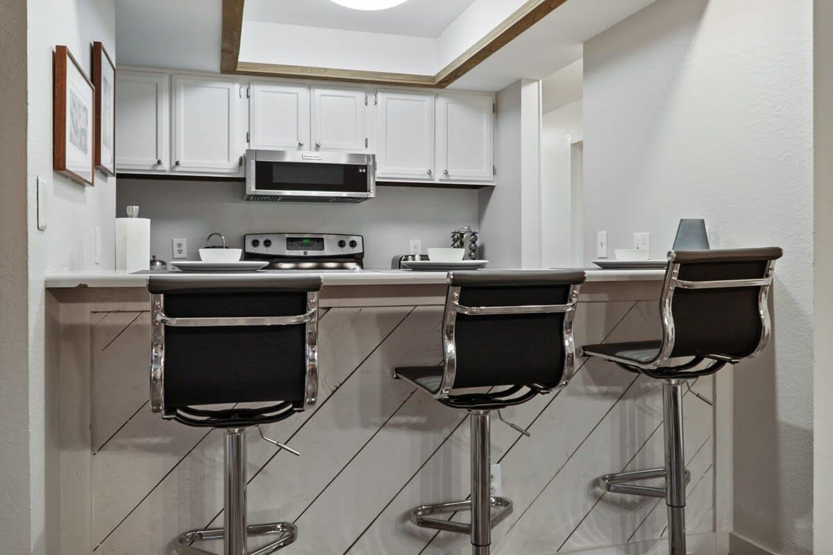 A modern kitchen with white cabinets, a microwave, a digital clock, and three bar stools.