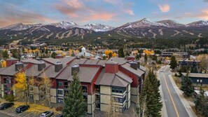 A residential area with a prominent building featuring a red and beige facade, a parking lot, and a road leading towards a mountainous backdrop.