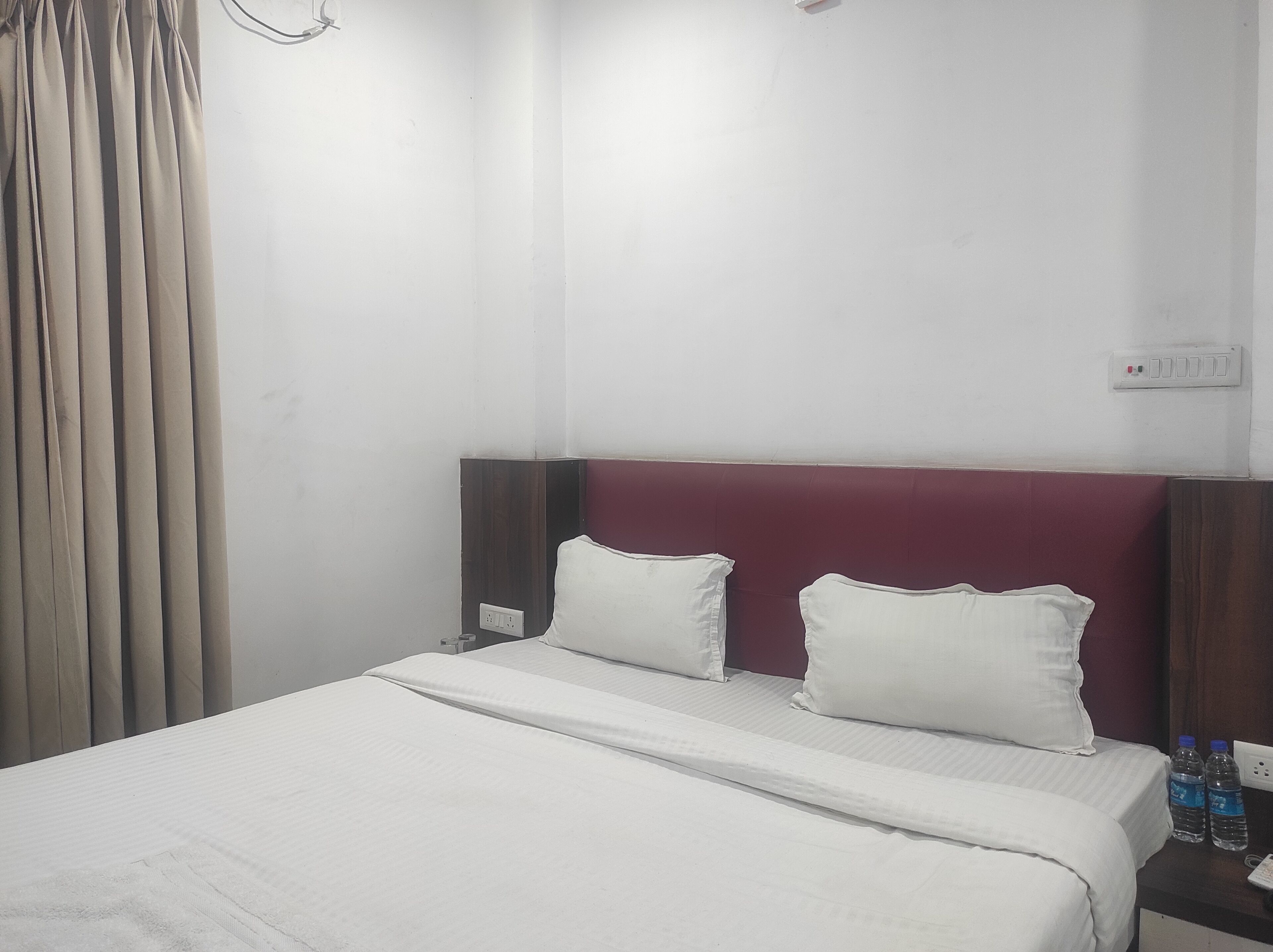 A hotel room with a bed, two pillows, a nightstand with water bottles, and a power outlet.