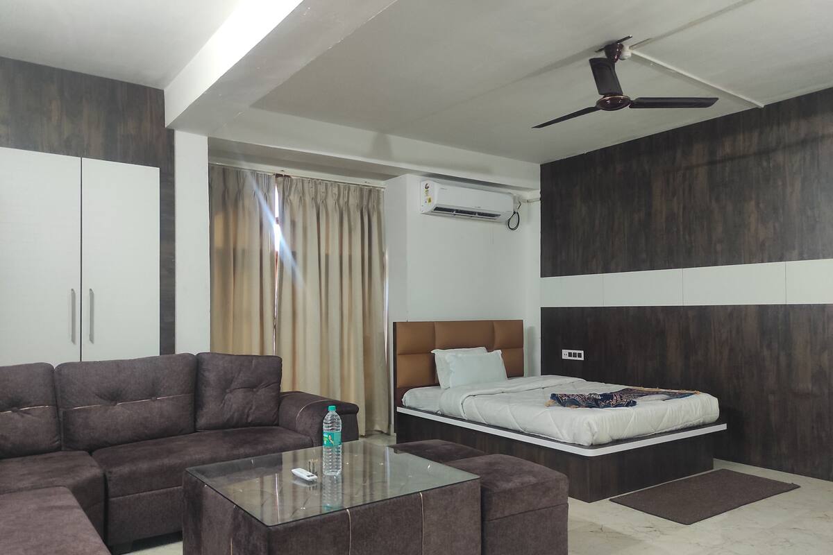 A modern hotel room with a sofa, a glass coffee table, a bed with a headboard, a ceiling fan, and a window with curtains.