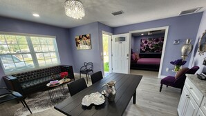 Uma sala de jantar com uma mesa de madeira escura, cadeiras pretas e um sofá preto. Há uma janela com persianas, uma porta que dá para outro cômodo e um vaso com flores na mesa.