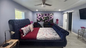 Quarto com uma cama grande, ventoinha no teto, janela com persianas, televisão fixada na parede e uma porta.