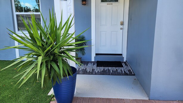 Uma casa com porta branca, placa de 'Não Perturbe', tapete de boas-vindas e um vaso azul grande com planta no jardim da frente.