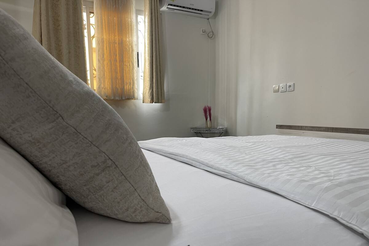 A hotel room with a bed, a window with curtains, an air conditioning unit, and a bedside table with a vase of flowers.