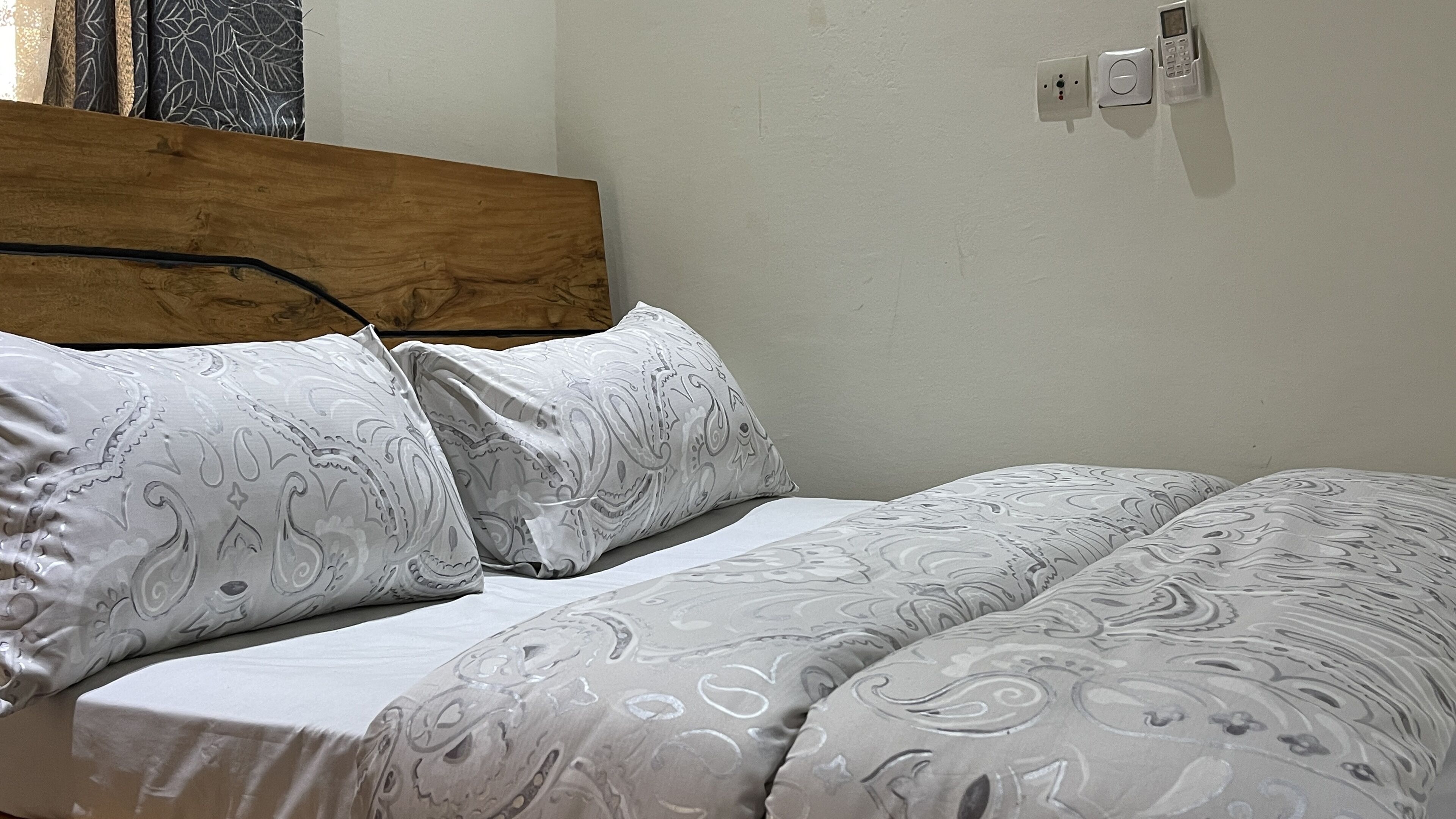 A bed with patterned bedding, a wooden headboard, and a wall-mounted light switch.