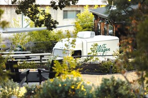 Ein weißer Wohnmobil mit der Aufschrift "The Wagon", geparkt in einem Gartenbereich mit Picknicktischen und einem Gebäude im Hintergrund.