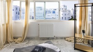 Un dormitorio moderno con un ventanal grande, un radiador, una cama con colcha a cuadros blanco y negro, y vistas a edificios.
