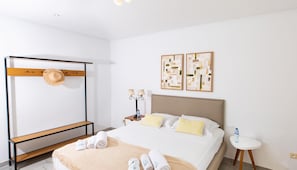 A hotel room with a bed, a bench, a hat rack, towels, a bedside table with a lamp, and two wall art pieces.