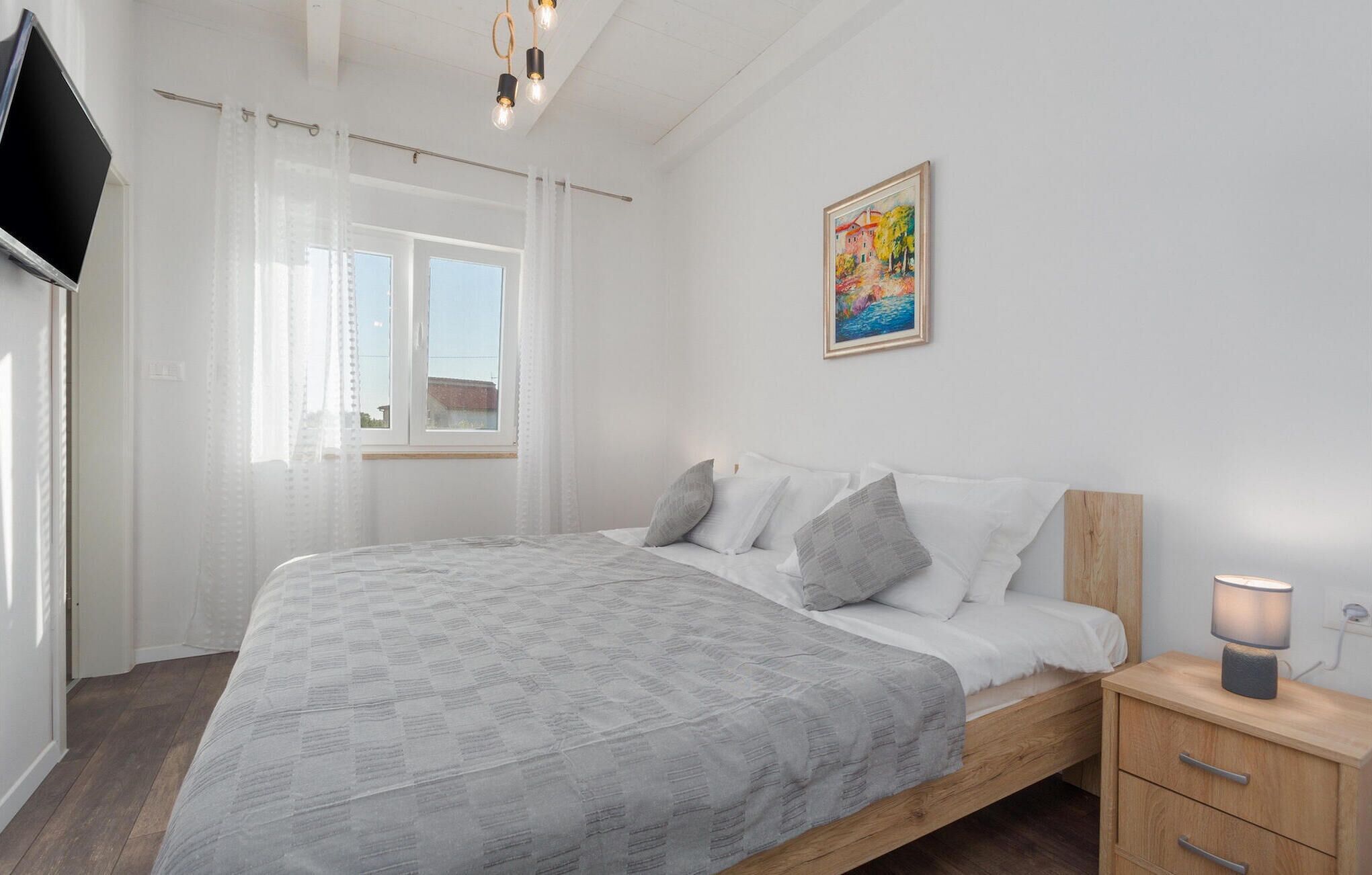 A bedroom with a bed, a bedside table, a lamp, a painting, and a window with curtains.