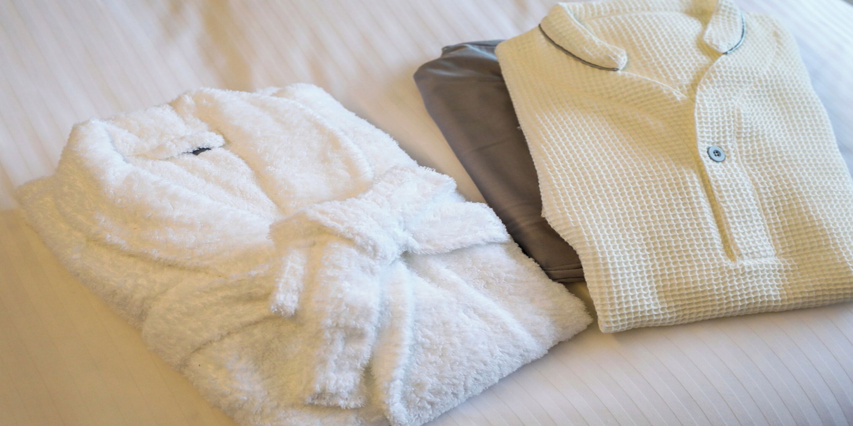A neatly folded white bathrobe and a white button-up shirt on a bed with a striped pattern.