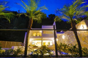 A two-story modern house with a balcony, surrounded by palm trees and a stone wall.