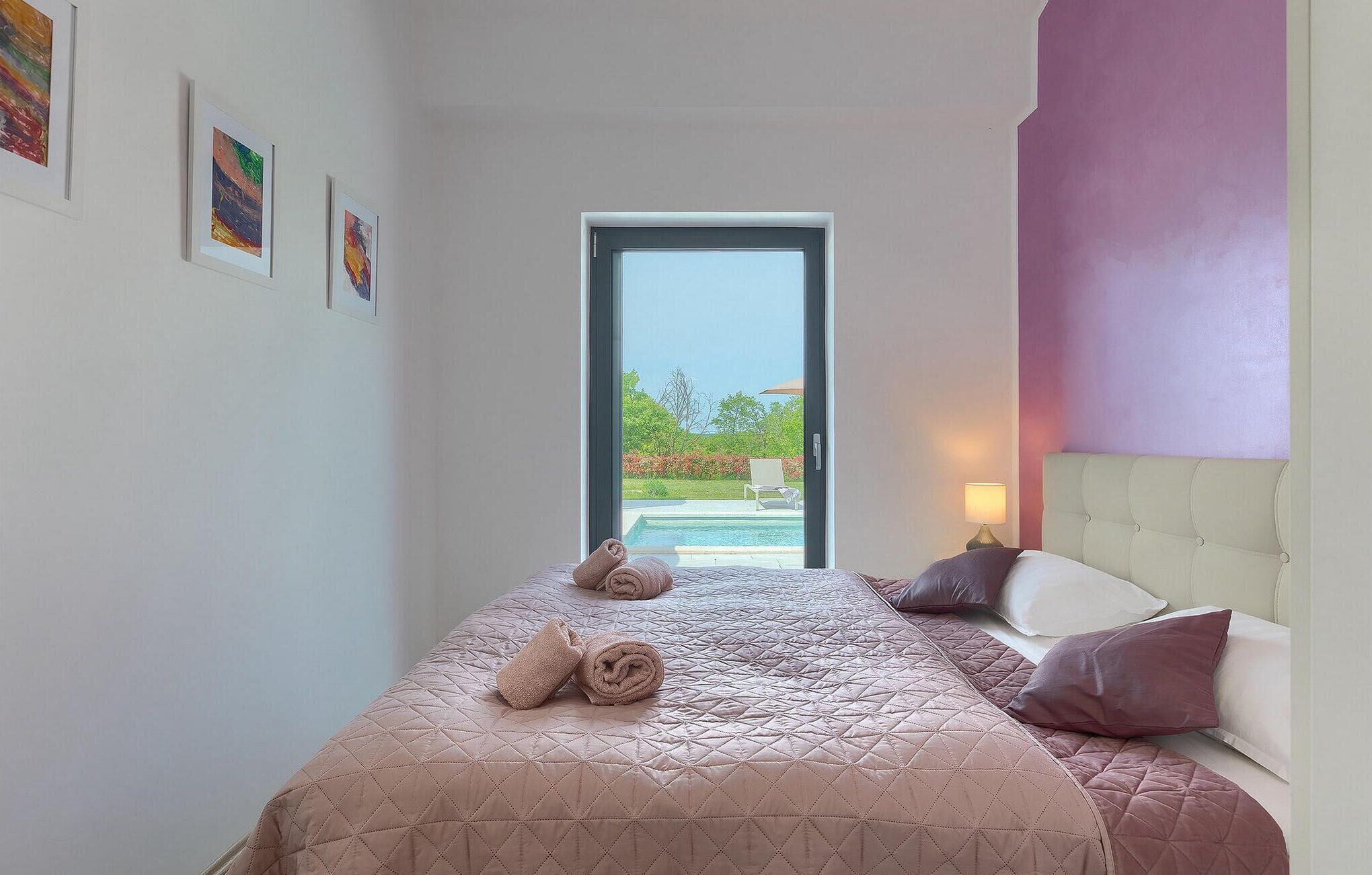 A hotel room with a bed, a nightstand, a lamp, and a view of a pool and outdoor seating.