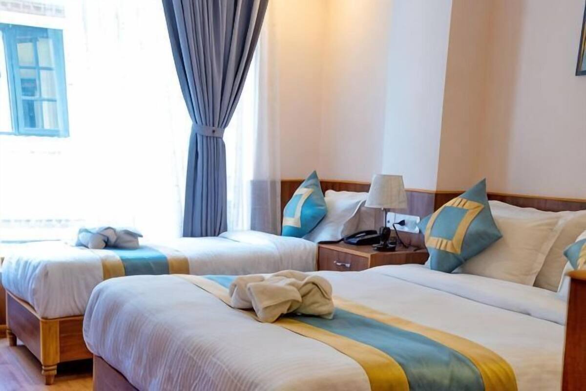 A hotel room with two beds, a nightstand, a lamp, and a window with curtains.