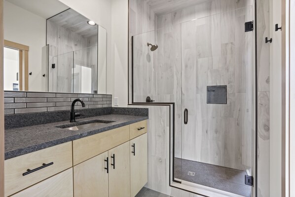 A modern bathroom with a granite countertop, a black faucet, and a glass-enclosed shower. - CAMP RANDONNEE__CABIN 4 (Mosier)