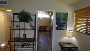 A hallway with a wooden floor, a metal bench, a table with a lamp, a plant shelf, and a painting on the wall. - Great conveniently located home. Near mall and many restaurants.  (Tucson)