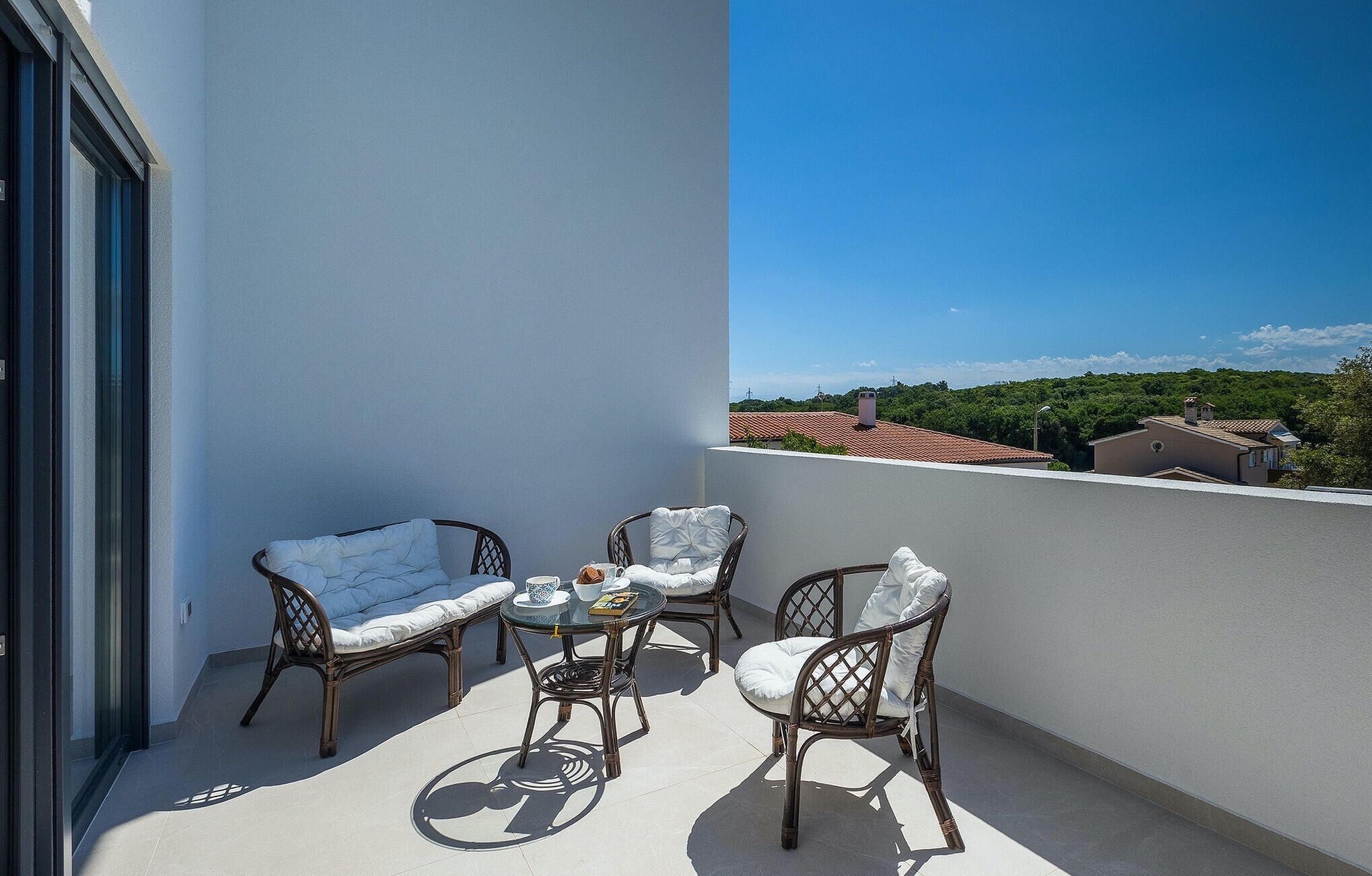 Ein Balkon mit Korbmöbeln, einem kleinen Tisch und Blick auf ein rotes Ziegeldach sowie grüne Bepflanzung im Hintergrund.